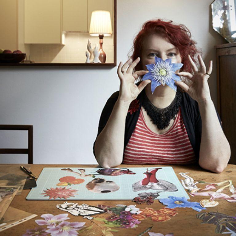 Deborah Kelly sits at a table with a collage in progress holding a paper flower in front of her face. 