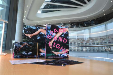 On a wooden desk overlooking a two-storeyed library filled with floor-to-ceiling-bookshelves, stands a collection of books. The front cover of these books depict a woman's legs floating against a backdrop of collaged sky and trees. Pink text curves around the cover, stating 'Space and Light'