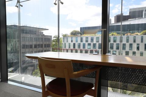 A wooden chair and desk with a white lamp faces a large floor-to-ceiling window. On the glass above the edge where the desk meets the glass, pink text dances across, stating, 'One feels over-exposed, violently lit up'