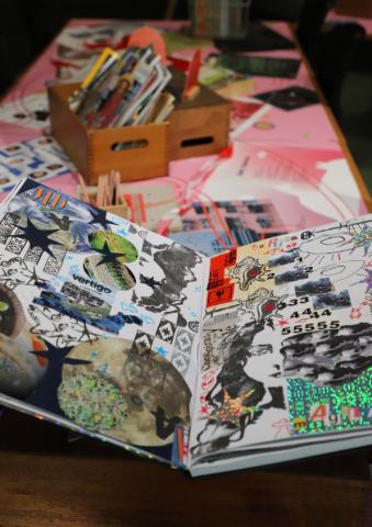 A large scrapbook filled with collages sits on top of a pink table, surrounded by craft supplies